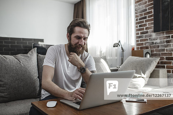 Lächelnder Mann sitzt auf der Couch zu Hause mit Laptop