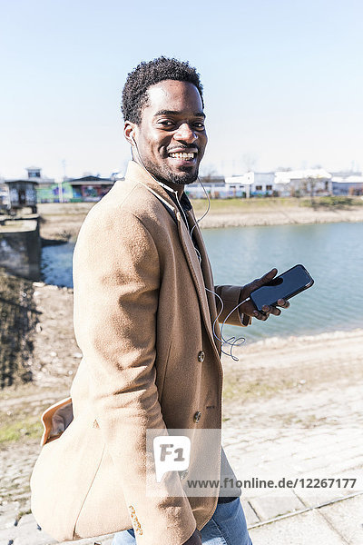 Porträt des lächelnden Geschäftsmannes am Wasser mit Handy und Kopfhörer