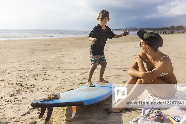 Indonesien  Bali  Junge auf dem Surfbrett stehend  Mutter am Strand sitzend