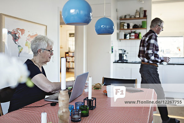 Seitenansicht der älteren Frau  die mit Laptop am Esstisch sitzt  während der Mann im Hintergrund läuft.