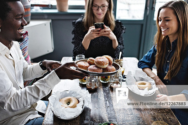 Smiling man giving buns to friend while woman using mobile phone at table