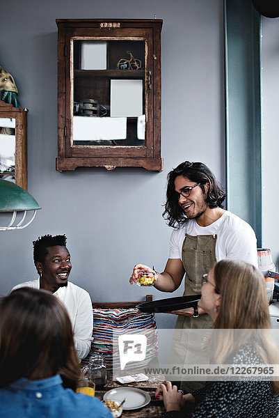 Lächelnder Besitzer serviert Essen für junge Männer und Frauen am Esstisch.