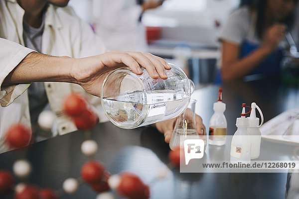 Mittelteil eines jungen männlichen Studenten  der flüssige Lösung in einen Kolben auf dem Tisch im Labor gießt.