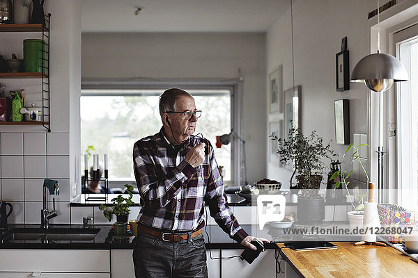 Senior man talking on smart phone through in-ear headphones while standing in kitchen at home
