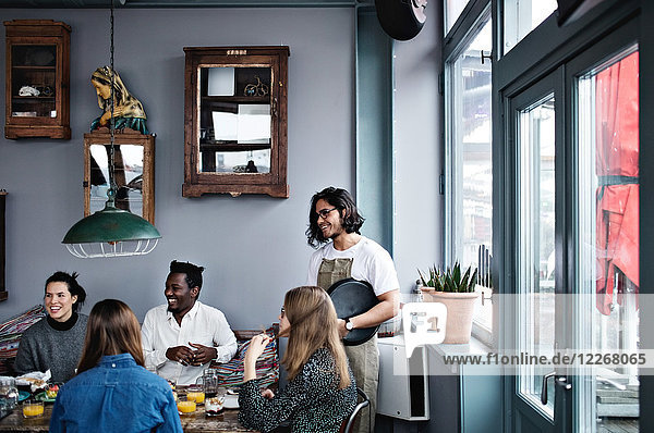 Smiling owner talking to young man and women at dining table in restaurant