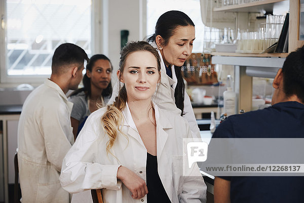 Portrait of confident young female student sitting against teacher friends in chemistry laboratory