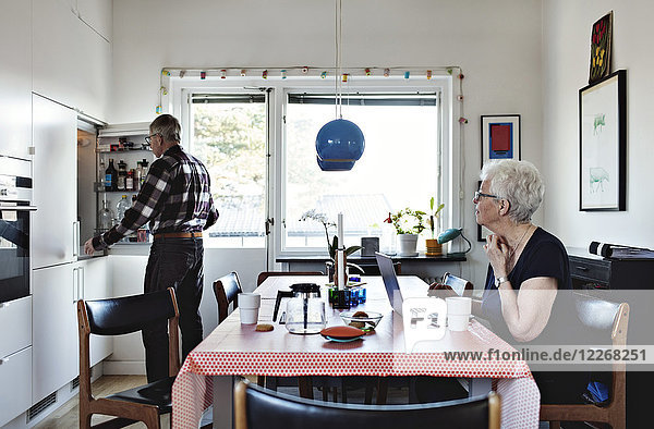 Seniorin sitzt am Esstisch  während der Mann am Kühlschrank in der Küche steht.