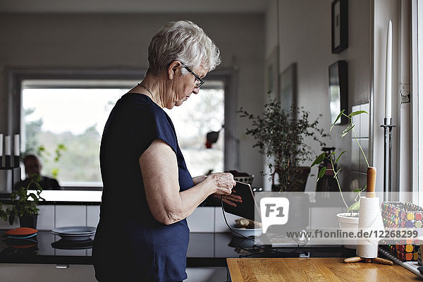 Seitenansicht der Seniorin beim Aufladen des digitalen Tabletts in der Küche zu Hause