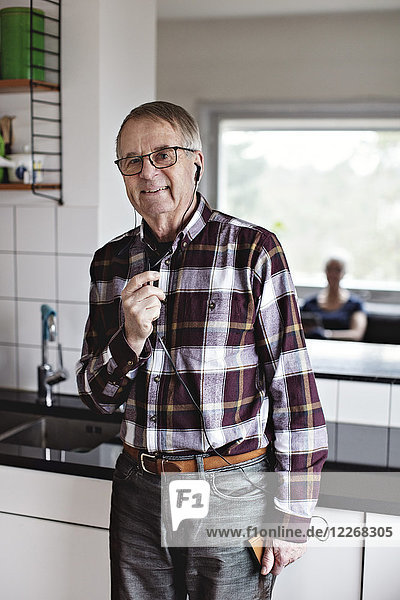 Porträt eines lächelnden älteren Mannes  der über In-Ear-Kopfhörer auf dem Smartphone spricht  während er zu Hause in der Küche steht.