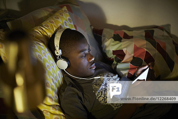 Hochwinkelansicht des Jungen  der Kopfhörer trägt und ein digitales Tablett benutzt  während er auf dem Bett liegt.