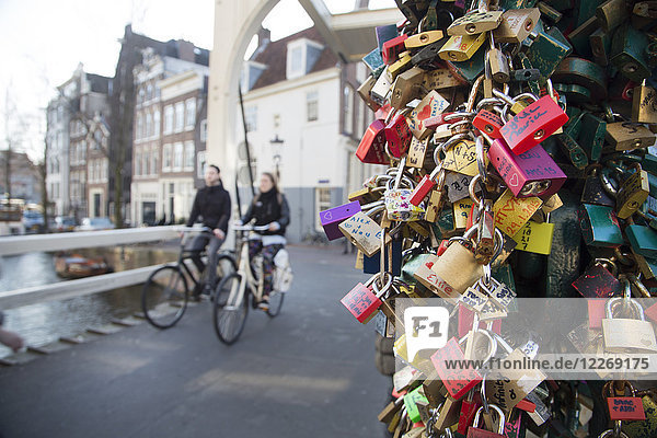 Nahaufnahme einer großen Zahl von Liebesvorhängeschlössern  zwei Personen auf Fahrrädern fahren entlang der Brücke über den Kanal in der Stadt.