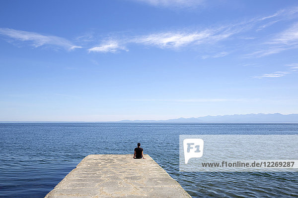 Rear view of person sitting on end of stone pier by the ocean.