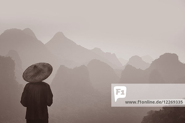 Rückansicht eines Mannes mit traditionellem Strohhut,  der über die Bergkette blickt.
