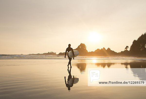 Reflexion eines Mannes  der einen Neoprenanzug trägt und bei Sonnenuntergang am Sandstrand spazieren geht und ein Surfbrett trägt.