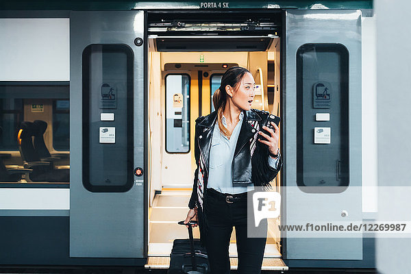 Frau steigt aus dem Zug  zieht Rollkoffer  hält Smartphone