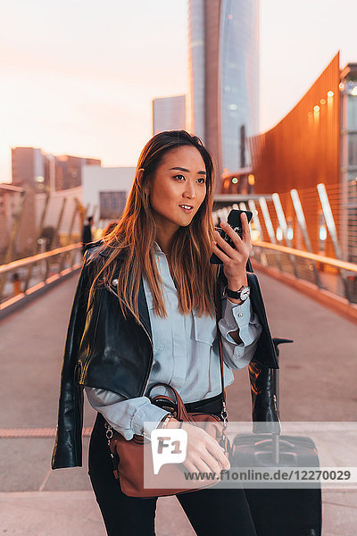 Junge Frau steht im Freien  benutzt ein Smartphone  neben ihr ein Koffer mit Rädern