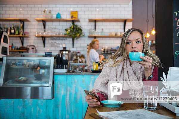 Frau sitzt im Café  hält ein Smartphone in der Hand und trinkt Kaffee