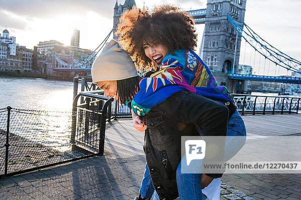 Young giving young woman piggyback outdoors  Tower Bridge in background  London  England  UK