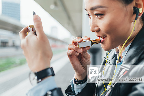 Woman sitting on train platform  applying lipstick  wearing earphones