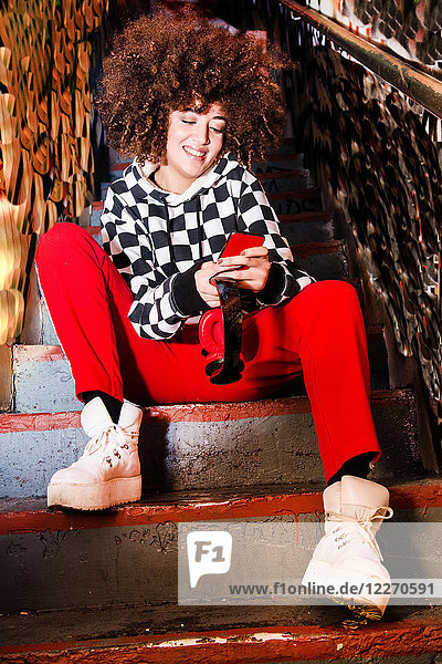 Junge Frau sitzt auf einer Treppe  benutzt ein Smartphone und lächelt