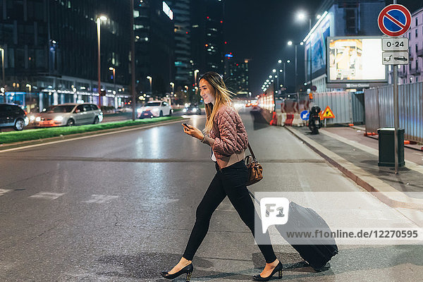 Woman walking outdoors at night  pulling wheeled suitcase  holding smartphone