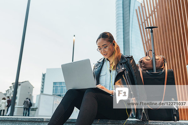 Businesswoman  sitting outdoors  using laptop  wheeled suitcase beside her