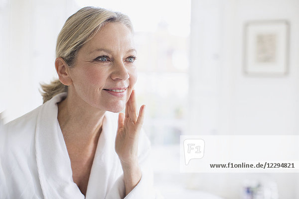 Smiling mature woman applying moisturizer to face at bathroom mirror