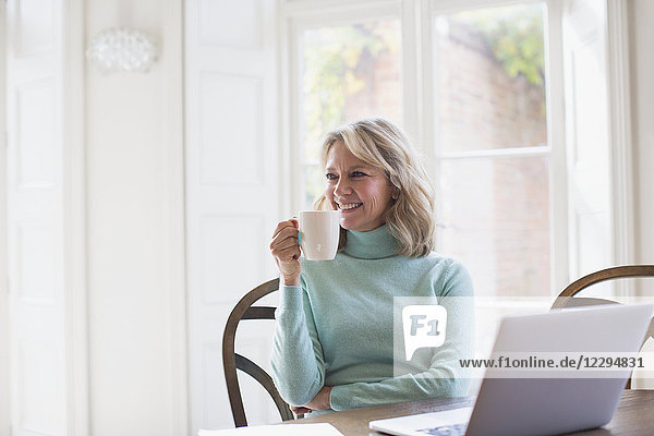 Lächelnde reife Freiberuflerin trinkt Tee und arbeitet am Laptop zu Hause