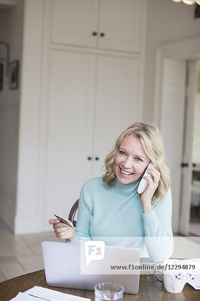 Lächelnde reife Frau mit Kreditkarte im Gespräch am Telefon am Laptop