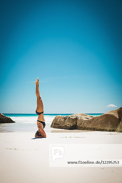 Mittlere erwachsene Frau im Bikini beim Kopfstand am Strand gegen den klaren blauen Himmel bei Sonnenschein