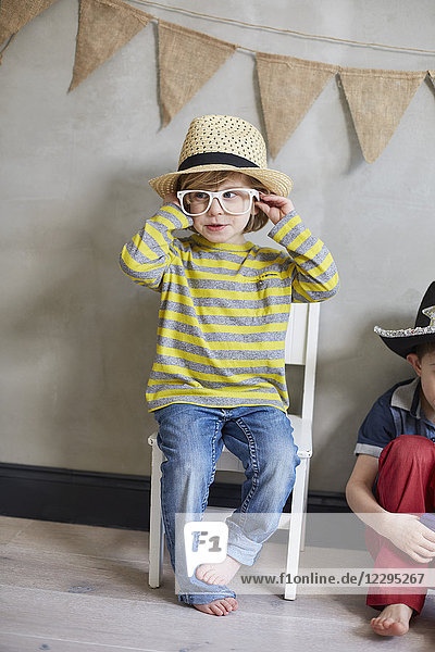 Glücklicher Junge mit Hut und Brille sitzt auf einem Stuhl von einem Freund gegen die Wand.