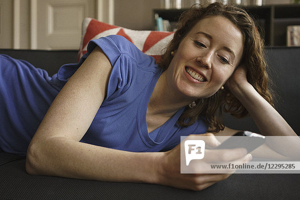 Lächelnde Frau auf dem Sofa liegend mit Smartphone zu Hause