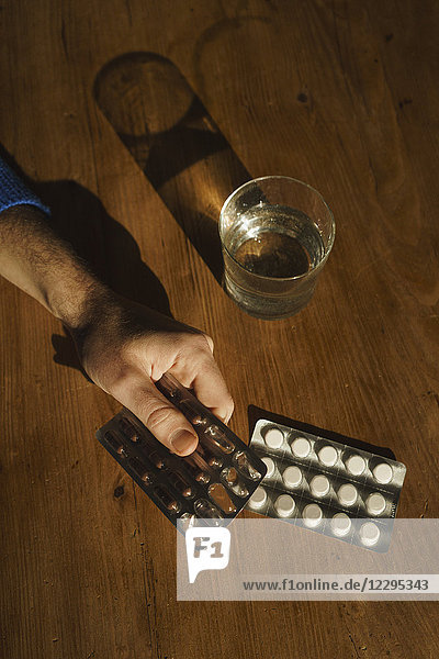 Abgeschnittene Hand des Mannes mit Medikamentenblisterpackung durch Trinkwasser auf dem Tisch zu Hause