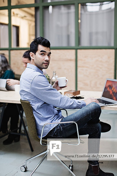 Portrait of businessman having coffee while using laptop at table in office