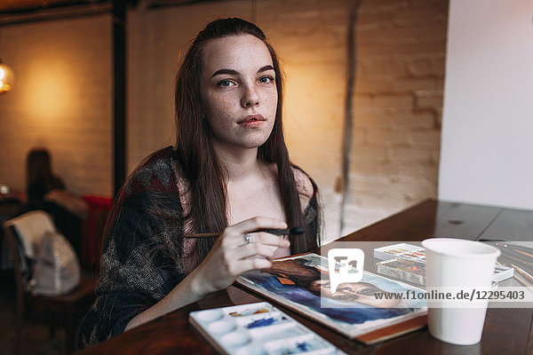 Portrait of beautiful young woman sitting by painting on table at cafe