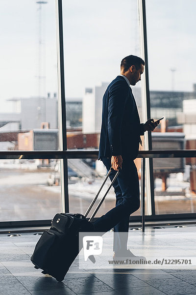 Full length side view of businessman with luggage using mobile phone while walking in airport