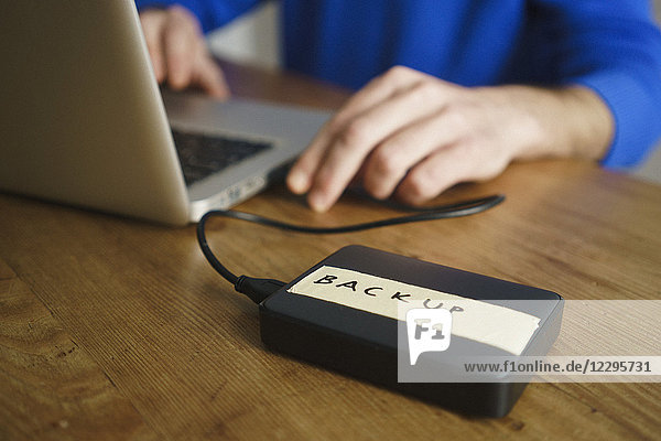 Midsection of man using laptop with backup label on external hard disk drive at table