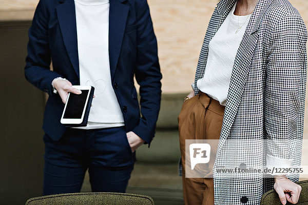 Midsection of businesswoman holding mobile phone while standing with female professional at office