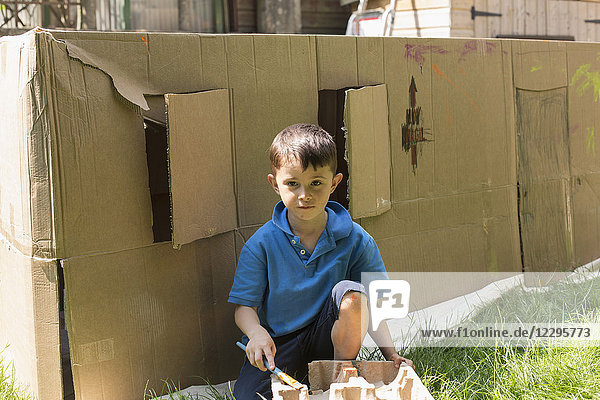 Porträt eines Jungen,  der an einem sonnigen Tag gegen ein Pappspielhaus im Hinterhof malt.