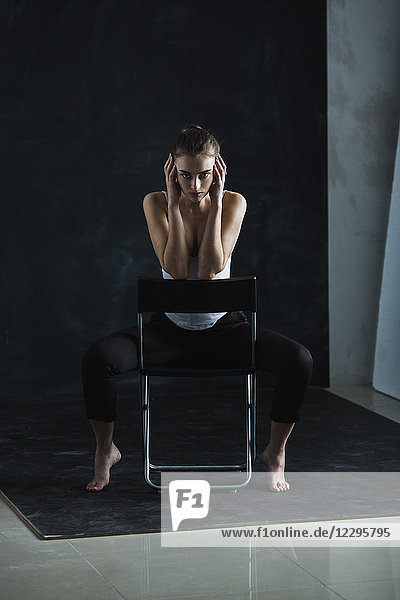 Full length portrait of young fashion model sitting on chair against black backdrop