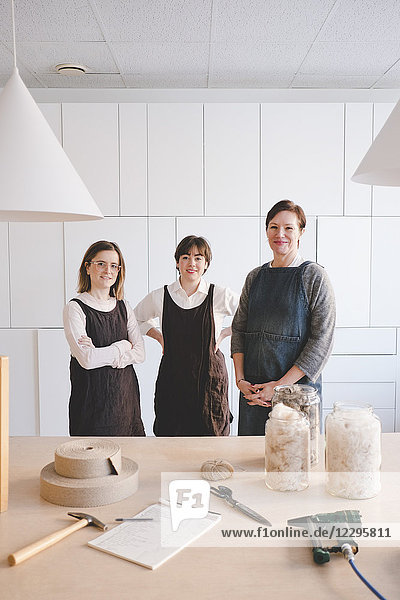 Portrait of confident design professionals wearing aprons with various equipment on desk in workshop