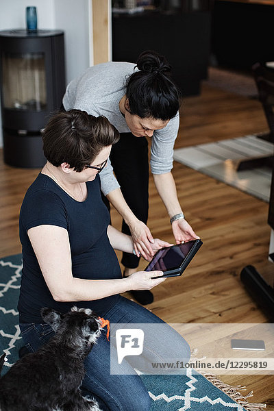 Woman showing digital tablet to girlfriend sitting by dog on carpet in living room