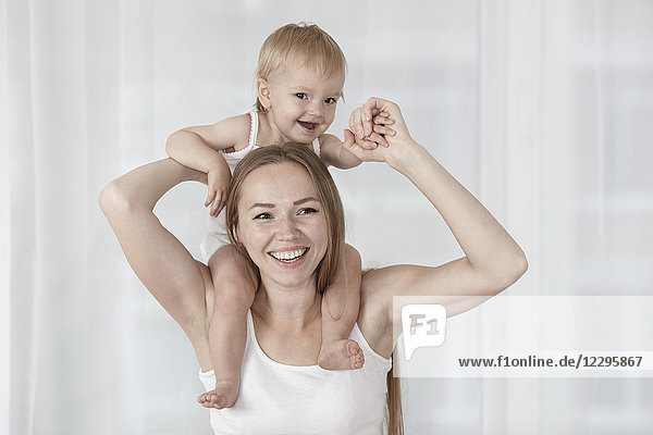Glückliche Mutter trägt Tochter auf Schultern gegen weißen Vorhang zu Hause