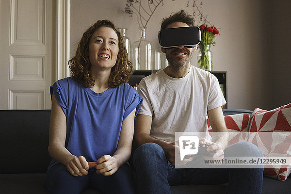 Smiling woman sitting by man playing on virtual reality headset in living room at home