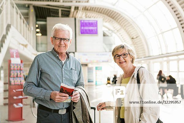 Portrait of smiling senior couple standing with book and luggage at subway station