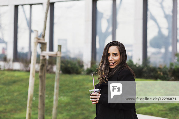 Happy mid adult woman holding drink while standing on grass against building