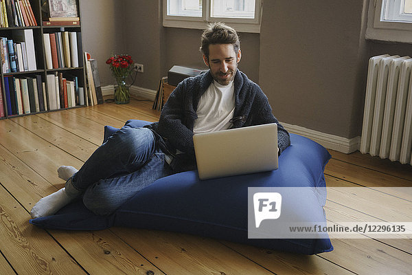 Volle Länge des Mannes sitzend mit Laptop auf blauer Bohnentüte im Zimmer zu Hause