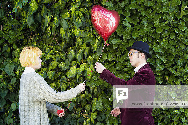 Side view of young couple holding heart shape balloon against plants at park