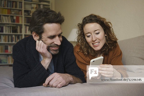 Smiling woman sharing smart phone with man lying on bed at home