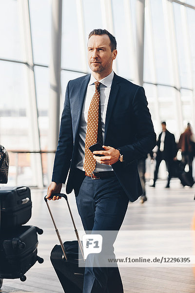 Confident mature businessman with mobile phone and luggage walking in airport